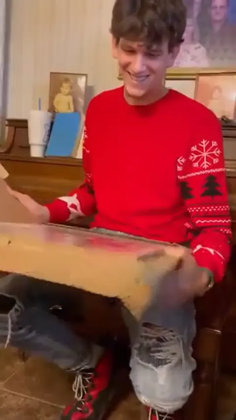 A young man in a red sweater smiling down at a portrait in its box.