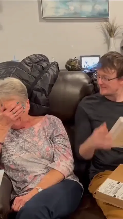 An older woman on a sofa, hand over her face, opening a portrait beside a young man.