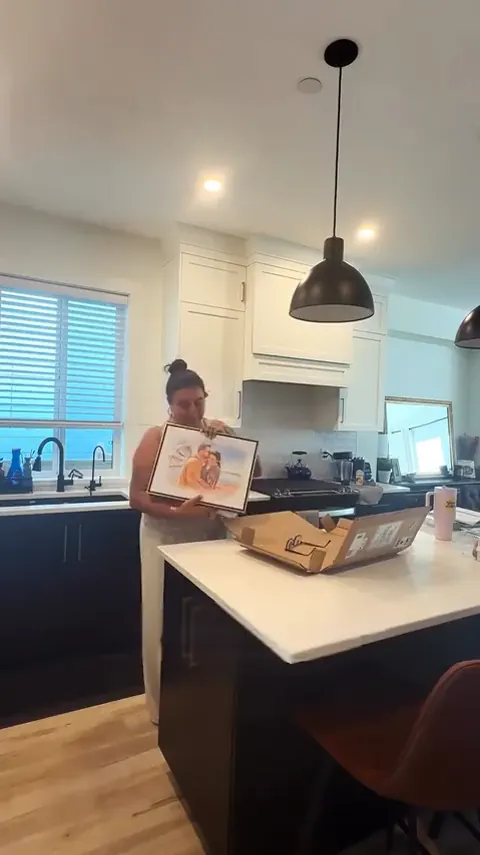 A woman in her kitchen holding up the framed portrait she has just unwrapped.